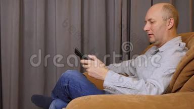 男人坐在扶手椅上，用遥控器在电视或音乐播放器上选择频道。